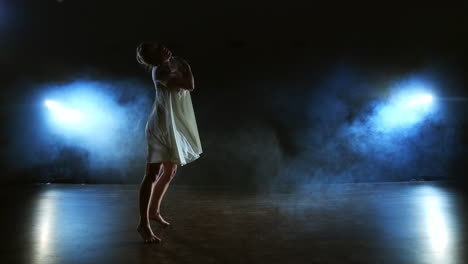 Ein-Modernes-Tanzmädchen-In-Einem-Weißen-Kleid-Tanzt-Ein-Modernes-Ballett-Und-Springt-Auf-Der-Bühne-Mit-Rauch-Im-Blauen-Scheinwerferlicht.